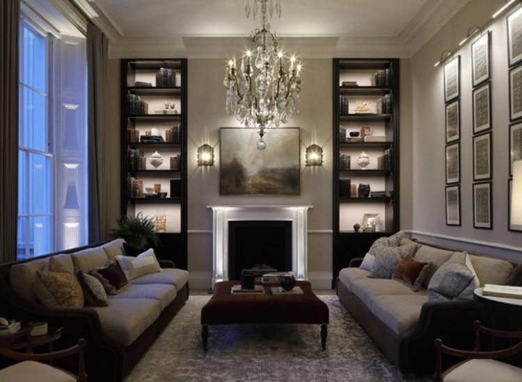 A Cozy Living Room With A Fireplace And An Elegant Chandelier And Indirect Lighting Of Shelves Illuminating The Space