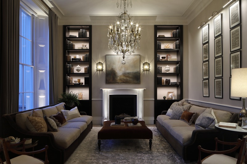 A cozy living room with a fireplace and an elegant chandelier and indirect lighting of shelves illuminating the space